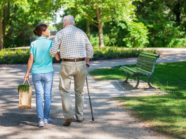 A caregiver carries groceries and accompanies an elderly man walking with a cane.