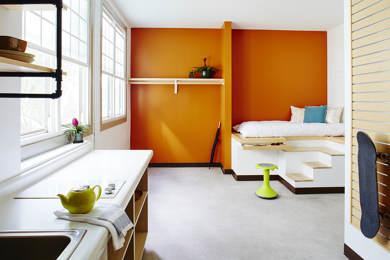 Photograph of the interior of an apartment, showing a small kitchenette, a bright orange wall, and a built-in platform bed. 