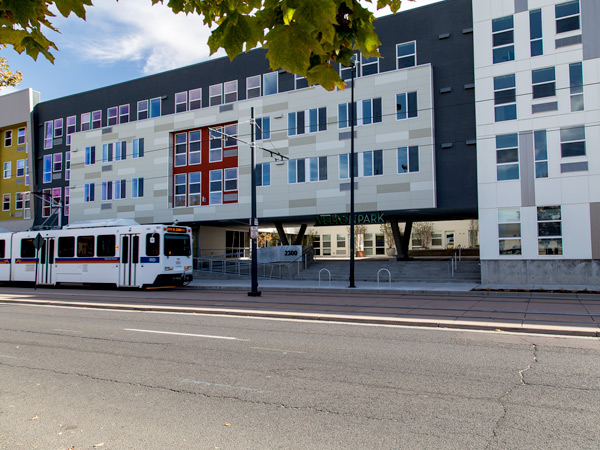 Photograph of the façade of a multi-story apartment building, with a light-rail train passing along a street in front. 