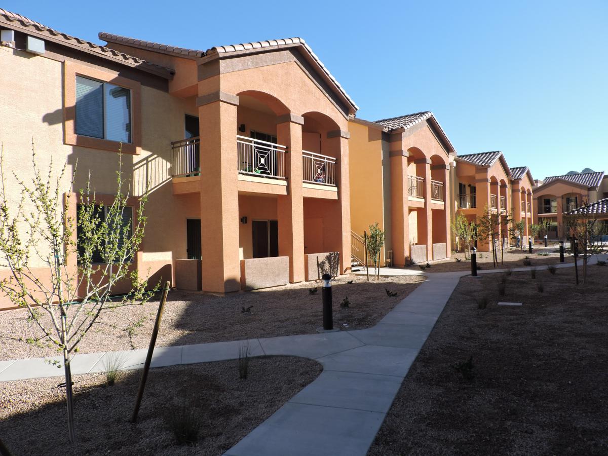 Exterior of two-story apartment buildings in Southwestern style facing sidewalks lined with young newly-planted trees.