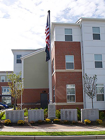 Five memorial stones with a flagpole in front of a three-story apartment building.