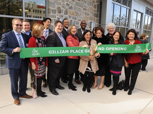 Photograph of group of people in front of a building holding a banner that says Gilliam Place Grand Opening being cut by one person in the center with scissors.