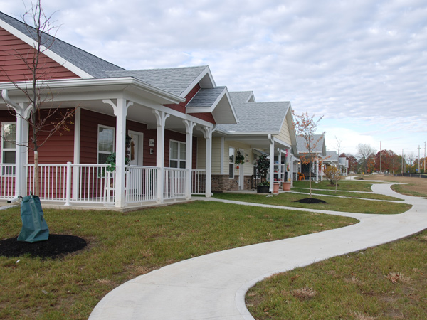 Photograph of a row of five single-story detached houses, each with a covered front porch facing out into open greenspace and connected by a winding path.