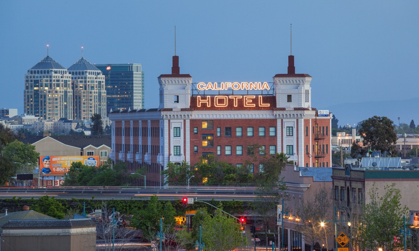 A photo of the front and side façade of a red brick and white stucco building topped by a marquee with a lit sign that reads “California Hotel.” A freeway is in the foreground.