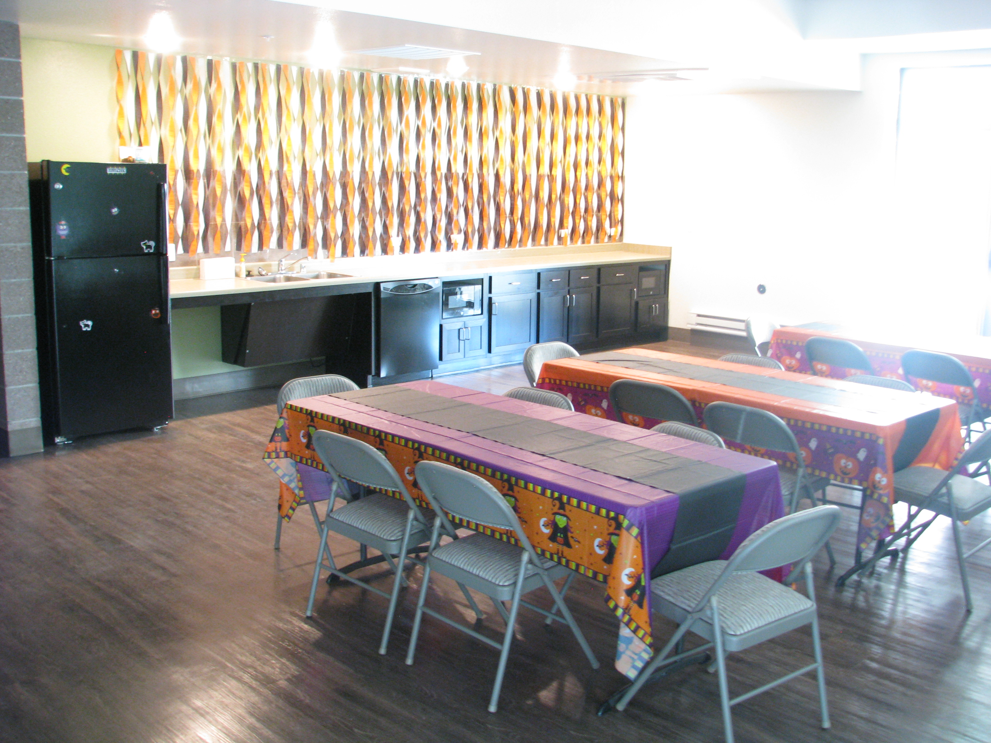 Photograph of a room with a kitchenette and several tables and chairs with Halloween-themed paper tablecloths. 