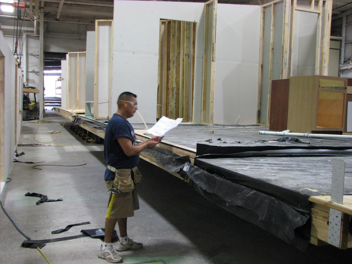 Photograph of a worker reading plans. Unfinished modular building components are visible in the background.