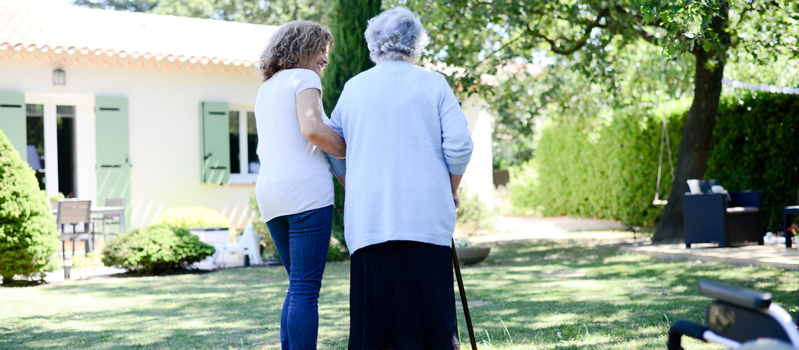 Photograph of a younger woman assisting an older woman walking with a cane in the backyard of a single-family home.