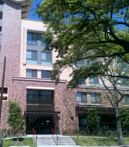 Photograph of the entrance to a five-story brick, metal, and vinyl-sided apartment building.