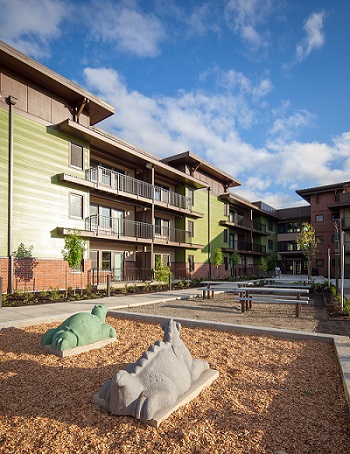 Photograph of a courtyard containing picnic tables, a play area, and landscaping, with a three-story apartment building in the background. 