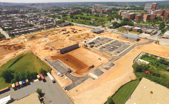 An aerial view of a large construction site and parking lot with buildings in the background.