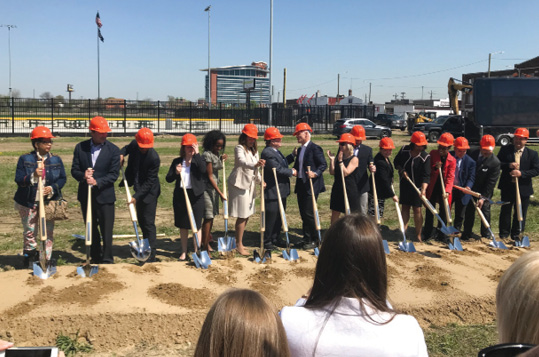 Photo shows 16 men and women wearing hardhats and holding shovels at a ground breaking ceremony.
