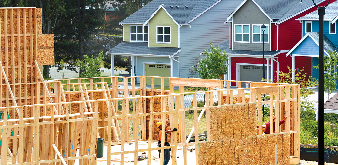Photo shows two workers framing walls for a house under construction with two completed houses in the background.
