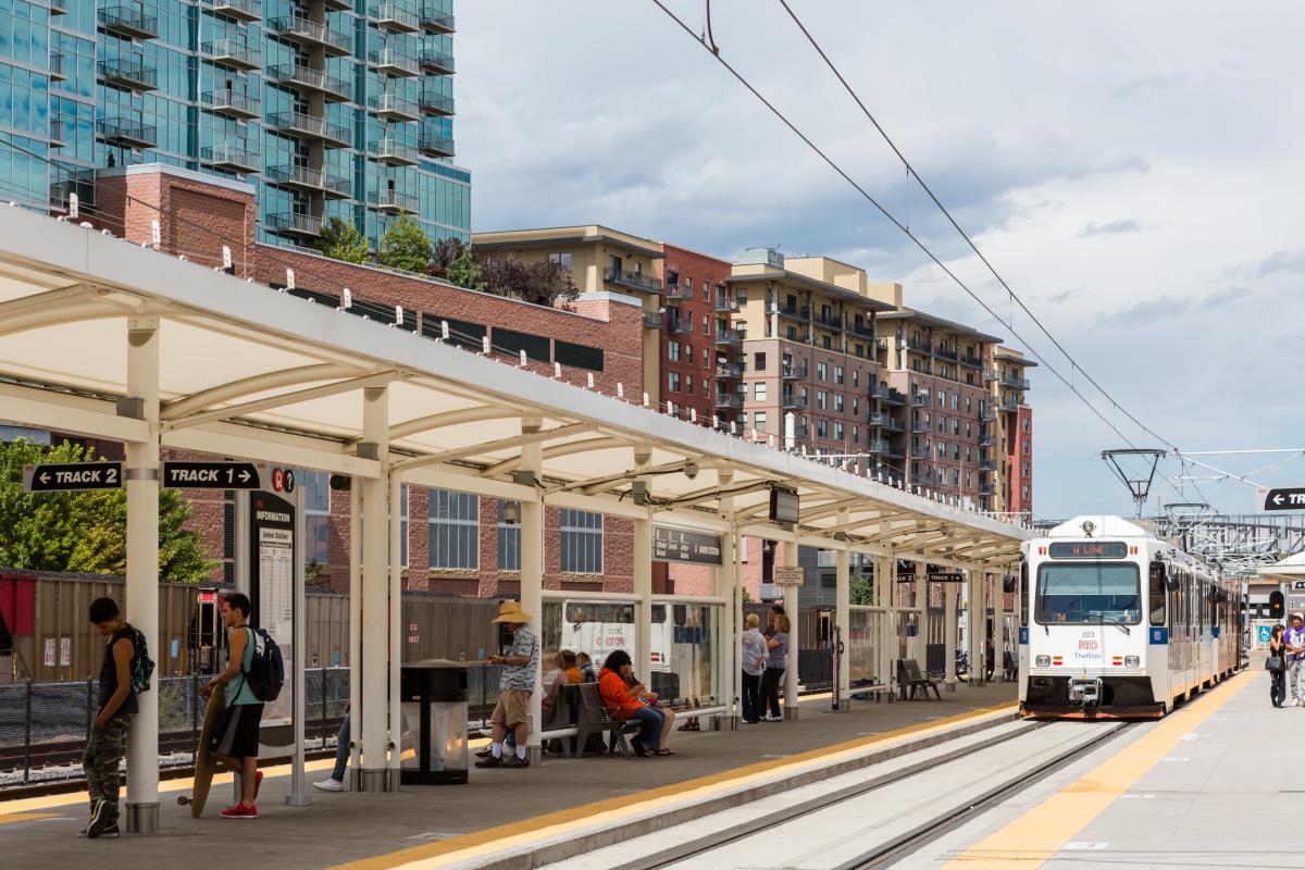 Photo shows the front of a light rail train along a covered platform with people and high rise buildings in the background.