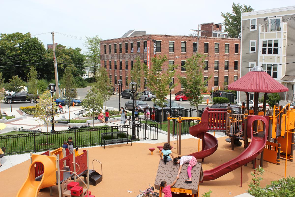 Children playing in an outdoor play area with a four-story and a three-story building in the background on the opposite side of the street.