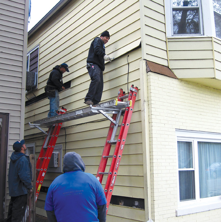 Two workers stand on a metal scaffolding plank working on the siding of a house as two others look on.