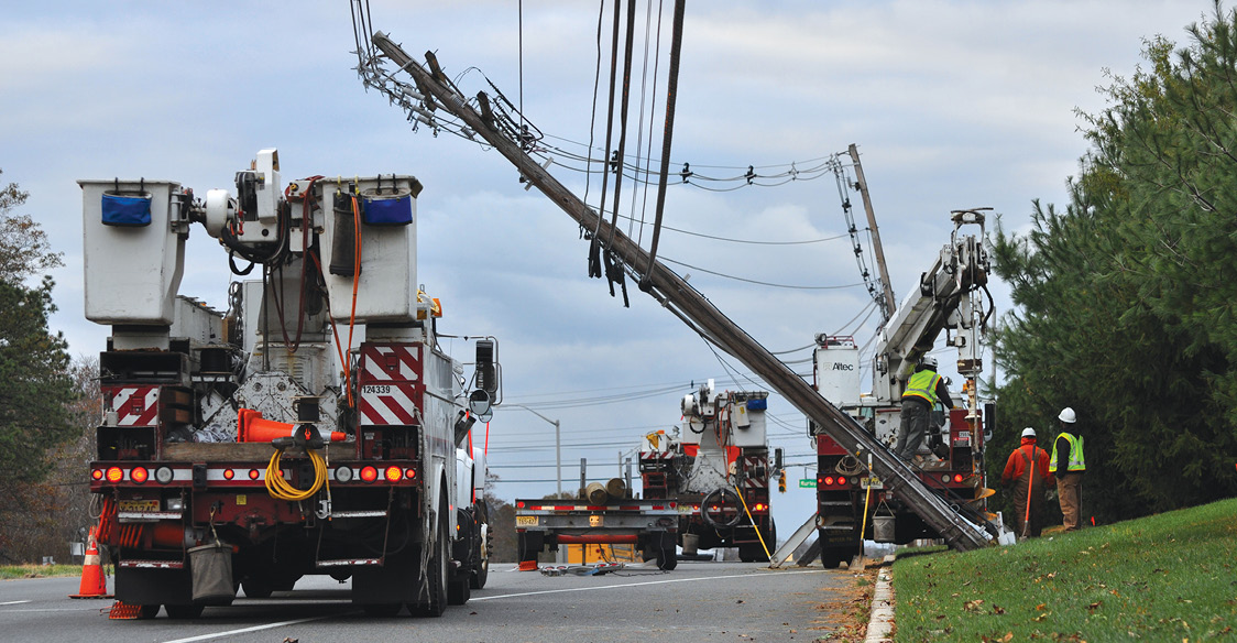 Photo shows three utility trucks and workers next to a utility pole leaning over a roadway.
