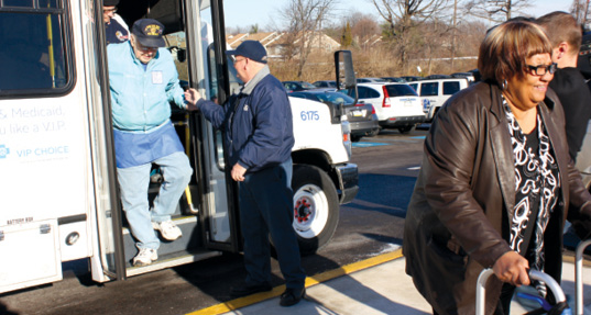 Photo shows an elderly man being helped off a van and an elderly woman with a walker in the foreground. 