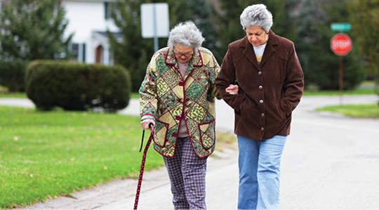 Photo shows two senior women, one of whom is using a cane, walking in a residential neighborhood. 