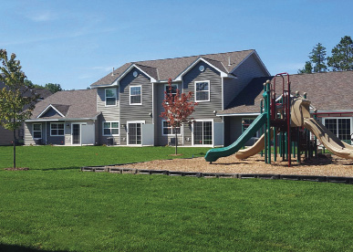 Photo shows one- and two-story attached homes with green lawn and a play area with slides in the foreground.