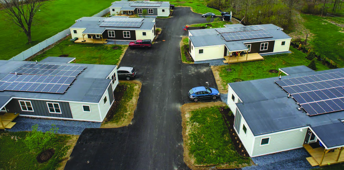 Low-angle aerial view of five modular homes located on both sides of a paved driveway with solar panels on the roofs.