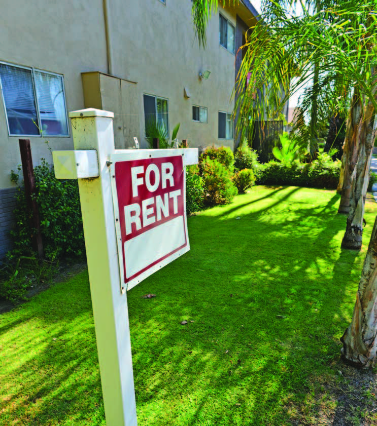 A “for rent” sign planted in grass in front of an apartment building.