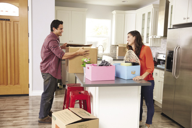 A man and woman stand on different sides of a kitchen island unpacking boxes in their home.