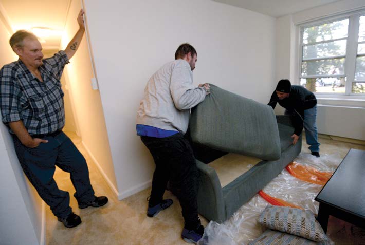 A man watches as movers assemble a sofa in his living room.