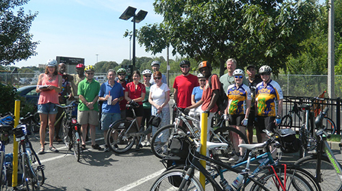 Photograph of 17 individuals, dressed in active apparel, standing next to several bicycles outside. 