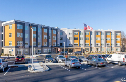 Photograph of a four-story residential building with a parking lot in front.