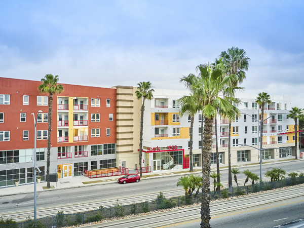  Aerial photograph of a five-story building along a street divided by a metro line with palm trees in the foreground. 