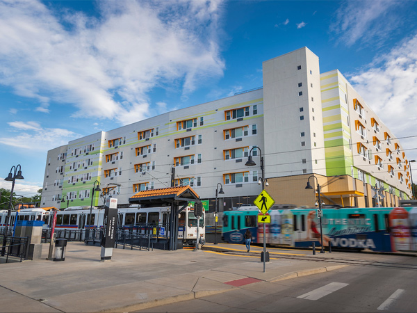 Photograph of the front and side facades of a four-story multifamily building, viewed across a street-level light rail station with two trains passing in opposite directions.