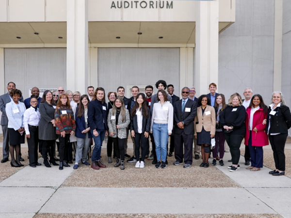 Group picture of 20-30 people outside in front of an auditorium.