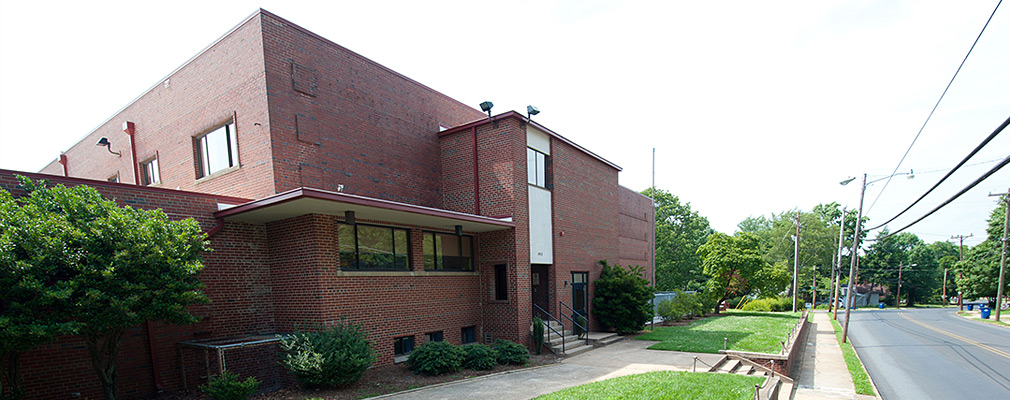 Photograph taken from street level of the front façade of a two-story, flat roofed building. The building, with the entrance in a bay projecting in front of the main façade, has few windows. The building is approximately three feet above the street on a terrace containing a narrow lawn, a walkway parallel to the building, and a line of shrubs directly in front of the façade.