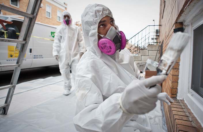 A man wearing protective gear is standing on a sidewalk painting a window frame. A man wearing protective gear is standing on a sidewalk painting a window frame.