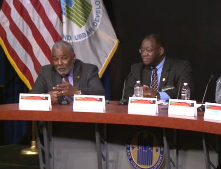 Image of two men seated at a table, with flags of the United States and HUD behind them.