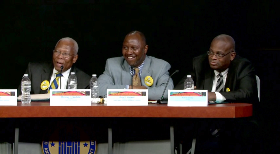 Image of three men seated at a table, with name plates and water bottles on the table and the HUD seal on the skirt of the table.