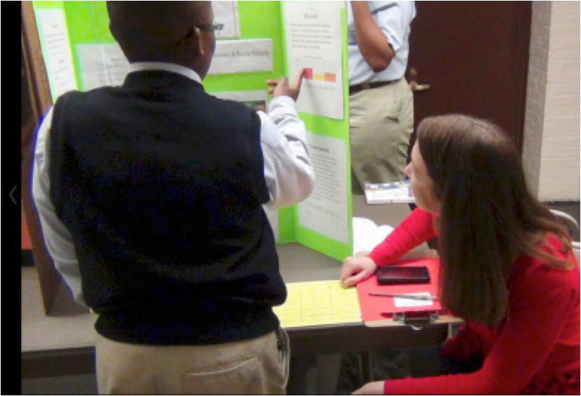 Photograph of an IIT professor standing before a display board in a meeting room, with a seated high school student listening to the professor’s comments.