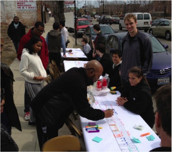 Photograph of approximately 10 adults looking at information on several tables manned by approximately seven IIT students. One resident is using a colored marker to provide comments on a diagram of commercial facades. The activity is taking place on a sidewalk of the street in the diagram. In the background are several commercial buildings and vehicles parked and moving in the street.