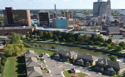 Photograph taken of downtown Flint, Michigan. Six low-lying buildings in the foreground lie adjacent to a river that runs through the center of the photograph. A variety of low- and mid-rise buildings can be seen in the background of the photograph.