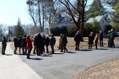 Image of over 20 individuals observing and photographing the Peter DeBaun House, partially obscured by shrubs and trees, and surrounding property.
