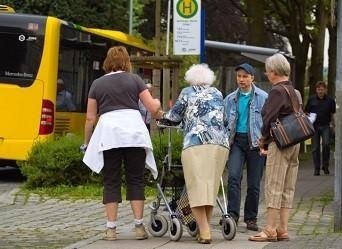 A senior woman with a walker and three other individuals gather at a bus stop, near a transit bus.