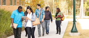 Photograph of six students carrying folders and tablets and walking on a sidewalk flanked by streetlights, vegetation, and buildings.