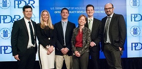 Photograph of six individuals, including five students from the New York University graduate student team and Assistant Secretary for Policy Development and Research Katherine O’Regan (third from the right), standing on stage in front of a PD&R banner.