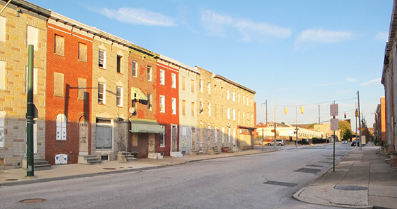 Exterior image of townhomes lining a street in East Baltimore. Boards cover the windows of several residential units.