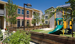 Photograph of a playground surrounded by a fence, small trees and shrubs, and low-rise apartments buildings.