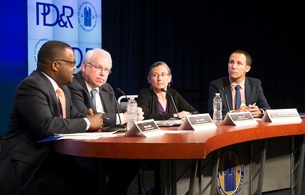 Four individuals sit at a table on stage with name cards and microphones; the PD&R logo is visible on a screen in the background.