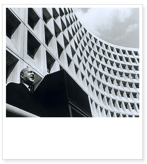 Robert C. Weaver, the first Secretary of HUD, speaks at a podium in front of the HUD building.