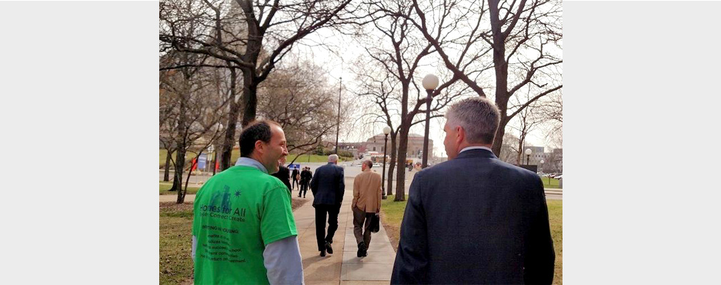 Two men, one wearing a Homes for All t-shirt, talking on a sidewalk.