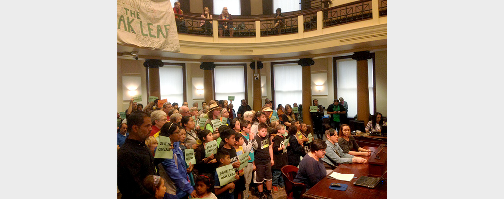 A group of children stand behind three women seated in front of microphones. 