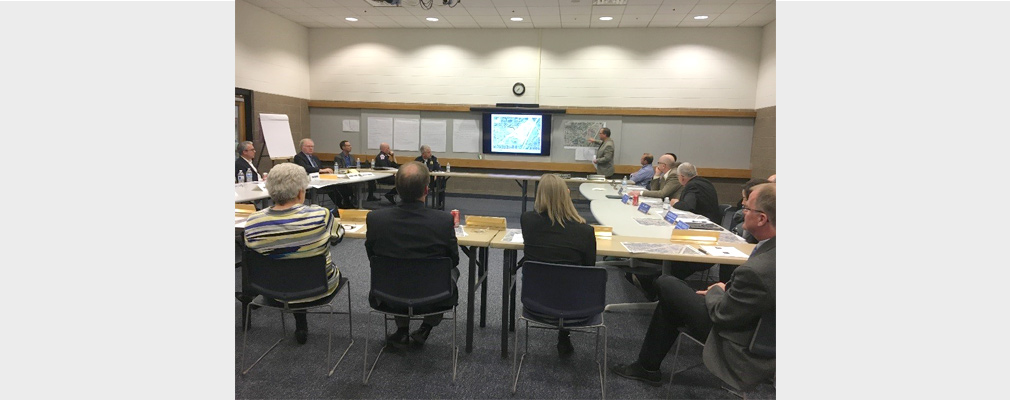 A group of men and women seated at tables arranged in a square listen to a presentation.